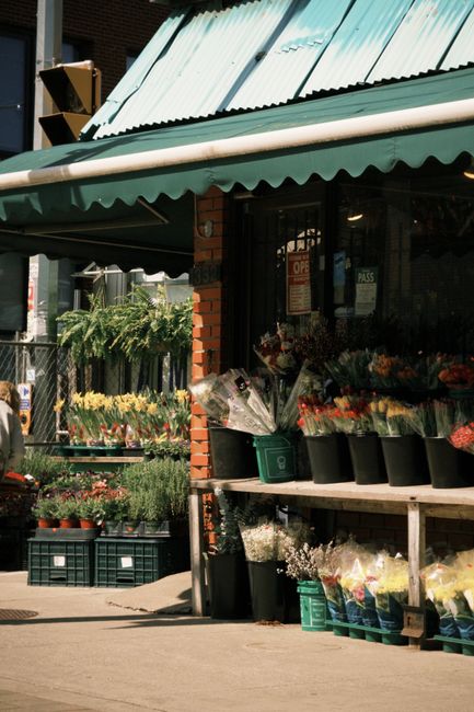 A display of fresh flowers outside a florist shop under a green awning, with various bouquets and potted plants arranged on wooden tables and black containers. The flowers include colorful roses, tuli