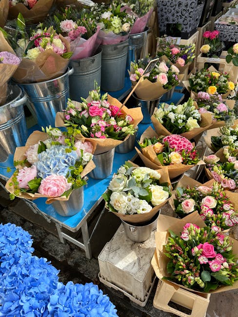 A vibrant display of freshly arranged flower bouquets on a blue table, featuring pastel pink and white roses, light blue hydrangeas, and assorted greenery wrapped in brown kraft paper. The bouquets ar