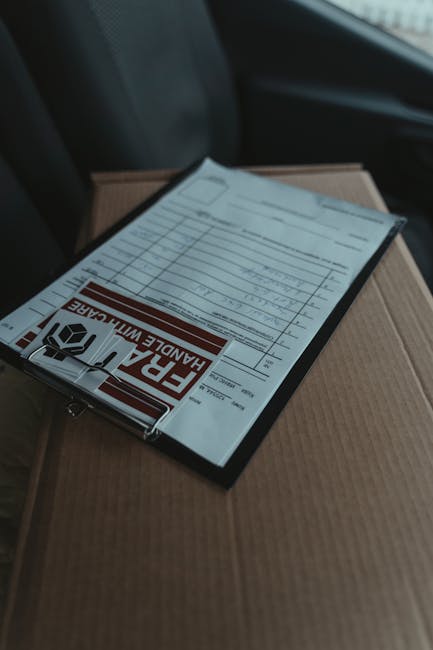 A close-up view of a cardboard box with a clipboard resting on top, containing a delivery receipt with a prominent red and black 'FRAGILE HANDLE WITH CARE' sticker. The background shows the interior o