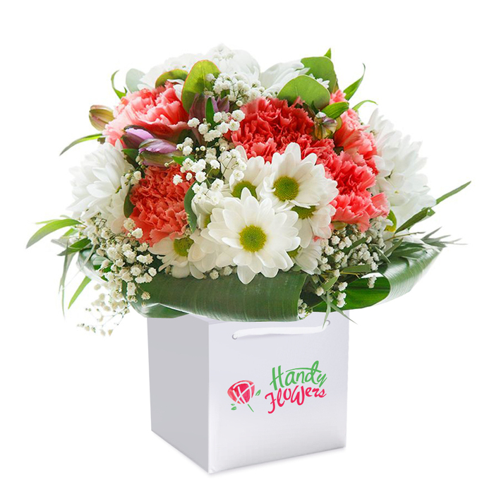 Set on a clean white backdrop, this exuberant bouquet reflects the refreshing character of Surrey Quays with its artful mix of flowers and foliage. Soft coral and vivid red carnations form the bouquet's core, their characteristic frilled petals spilling outwards and catching the light-an effect matched only by blossoms found in the parks near Surrey Water. Amid them, crisp white daisies with striking yellow-green centers provide cheerful highlights, punctuating the arrangement with a playful brightness reminiscent of early spring mornings at South Dock. Wisps of delicate baby's breath (gypsophila) are interspersed throughout, their lacy texture floating like London fog and softening the overall silhouette. Nestled discreetly among the open florals are a few closed buds with hints of purple and blush, adding subtle depth, intrigue, and a touch of anticipation for blooms yet to unfurl. Encircling the entire display, broad, shiny green leaves create a natural, elegant collar, holding the flowers upright and accentuating their color and beauty. The bouquet is thoughtfully composed and stands upright within a sharp, white square gift bag-its handles designed for both style and practicality, perfect for gifting or seamless delivery around the wharves or local cafes. No branding distracts from the scene; the focus remains on the florals and the skill with which they're arranged. Captured with professional clarity, this image evokes vibrancy, freshness, and a celebration of Surrey Quays' distinctive blend of community and nature-making it an ideal symbol for a birthday, anniversary, or any occasion deserving a locally crafted floral tribute.